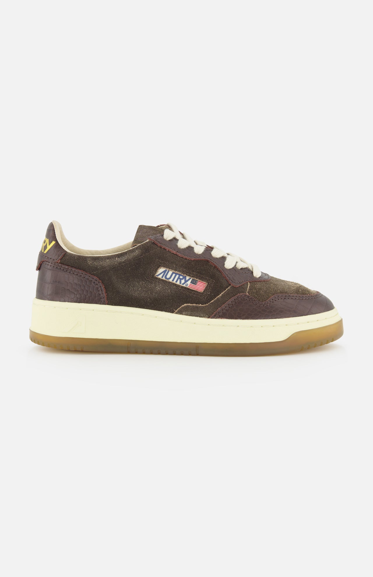 Introducing the AUTRY Medalist Low Suede Sneaker with Croco-effect: a low-top sneaker crafted from rich dark chocolate brown suede with distinctive croco-effect detailing. It features crisp white laces, a gum sole, and is finished with a small American flag logo on the side that reads AUTRY. Inspired by classic 1980s tennis shoes, this design effortlessly blends vintage appeal with contemporary style.
