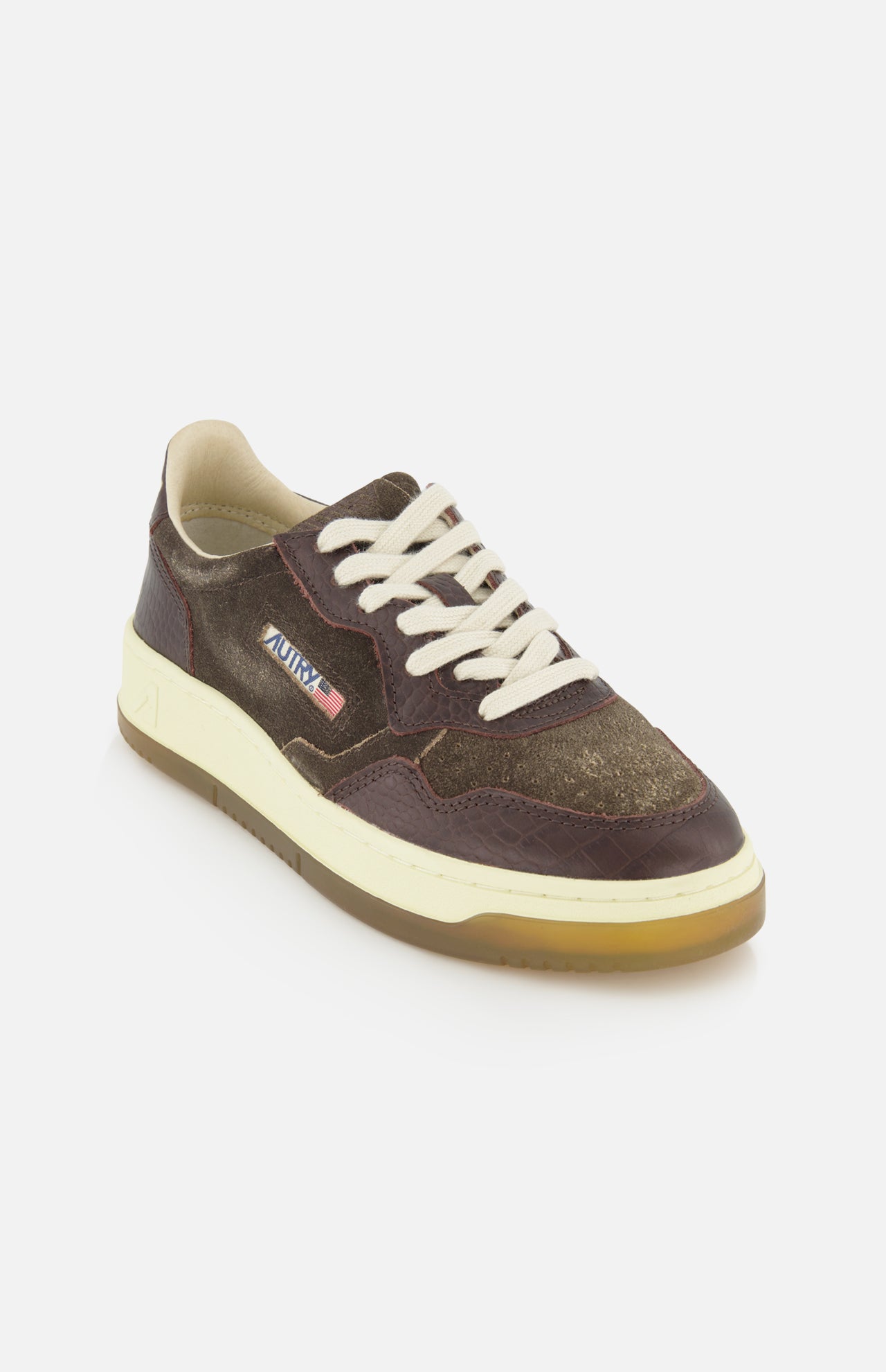 The AUTRY Medalist Low Suede Sneaker with Croco-effect is shown on a white background, featuring brown and olive tones, white laces, a beige sole, and a small American flag patch on the side. Its dark chocolate brown suede accents evoke the style of classic 1980s tennis shoes.