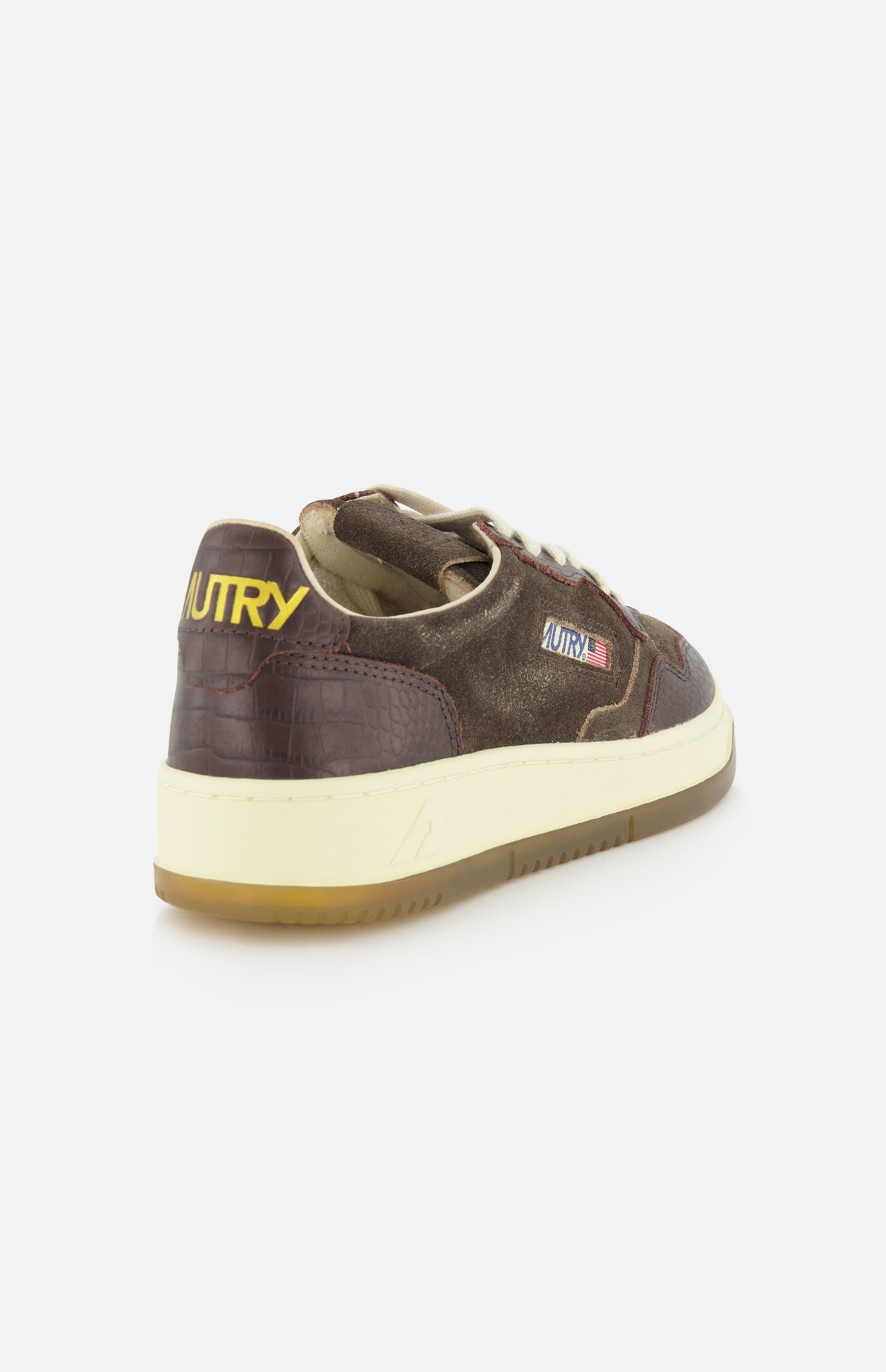 Introducing the AUTRY Medalist Low Suede Sneaker with Croco-effect: a brown low-top crafted from dark chocolate suede, boasting a refined crocodile-effect texture. This sneaker is highlighted by AUTRY’s signature yellow branding on the heel, a crisp white sole, and a tan gum outsole—capturing the authentic vintage style of 1980s tennis shoes when seen from the back angle.