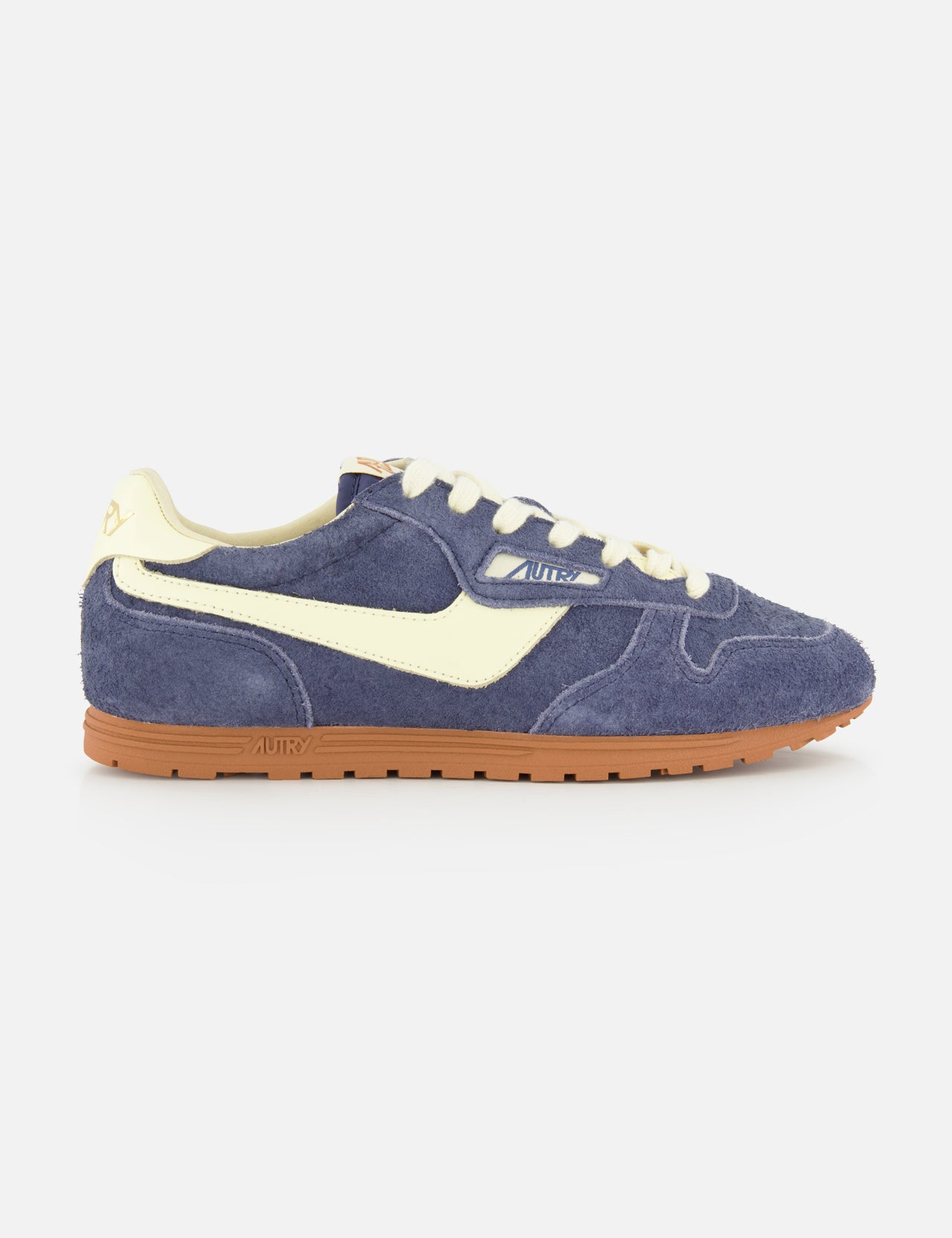 The AUTRY Windspin Low Suede Sneaker features dark blue suede with a creamy side stripe, laces, and heel tab, set atop a brown rubber sole. Photographed on a plain white background, this vintage-inspired design brings effortless 60s style to any modern wardrobe.