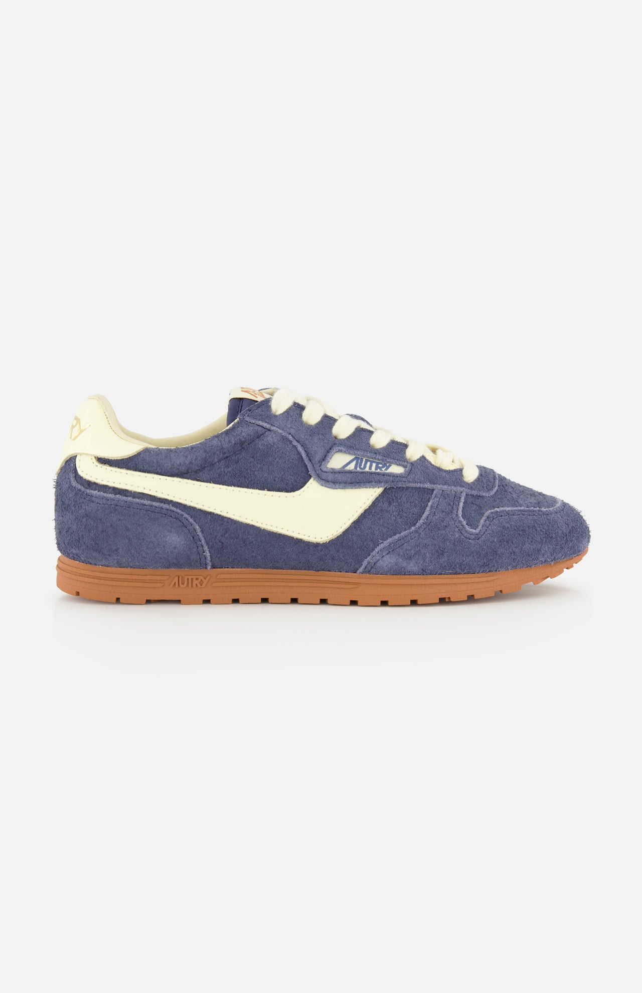 The AUTRY Windspin Low Suede Sneaker features dark blue suede with a creamy side stripe, laces, and heel tab, set atop a brown rubber sole. Photographed on a plain white background, this vintage-inspired design brings effortless 60s style to any modern wardrobe.