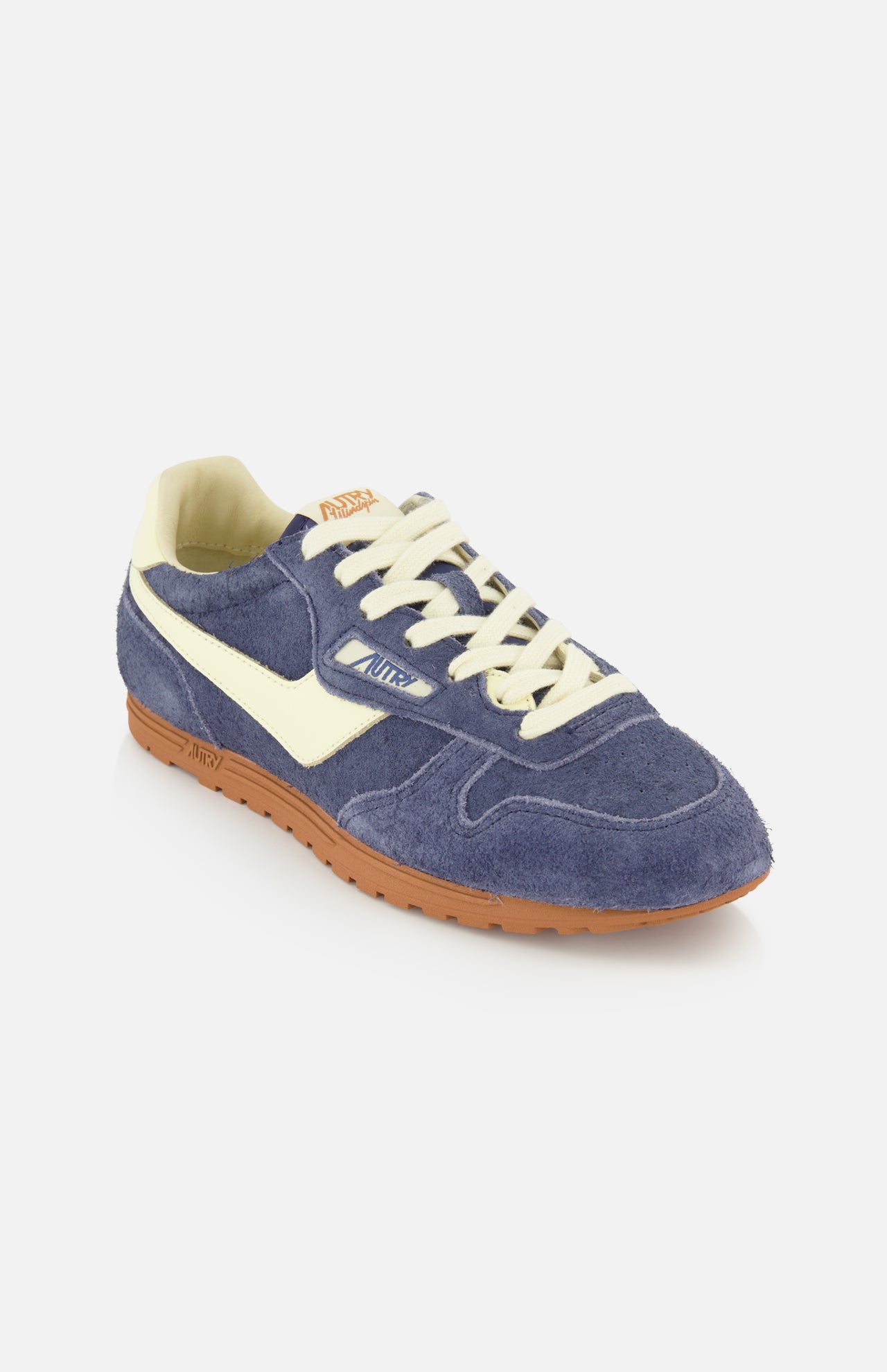Introducing the AUTRY Windspin Low Suede Sneaker, featuring dark blue suede with a soft textured finish, cream laces, a signature white side stripe, and a brown rubber sole—capturing retro-inspired charm against a clean white backdrop.