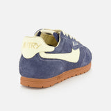 The AUTRY Windspin Low Suede Sneaker is showcased against a plain white background, featuring dark blue suede for vintage appeal, a cream heel tab with AUTRY branding, a cream side stripe, crisp white laces, and a brown rubber sole inspired by 60s style.