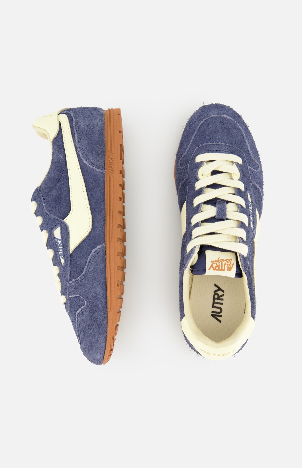 The AUTRY Windspin Low Suede Sneaker is showcased from the side and top against a white background, featuring a blue and cream retro-inspired design with gum soles. Its dark blue suede accents capture the timeless appeal of classic 60s-style sneakers, embodying vintage charm.