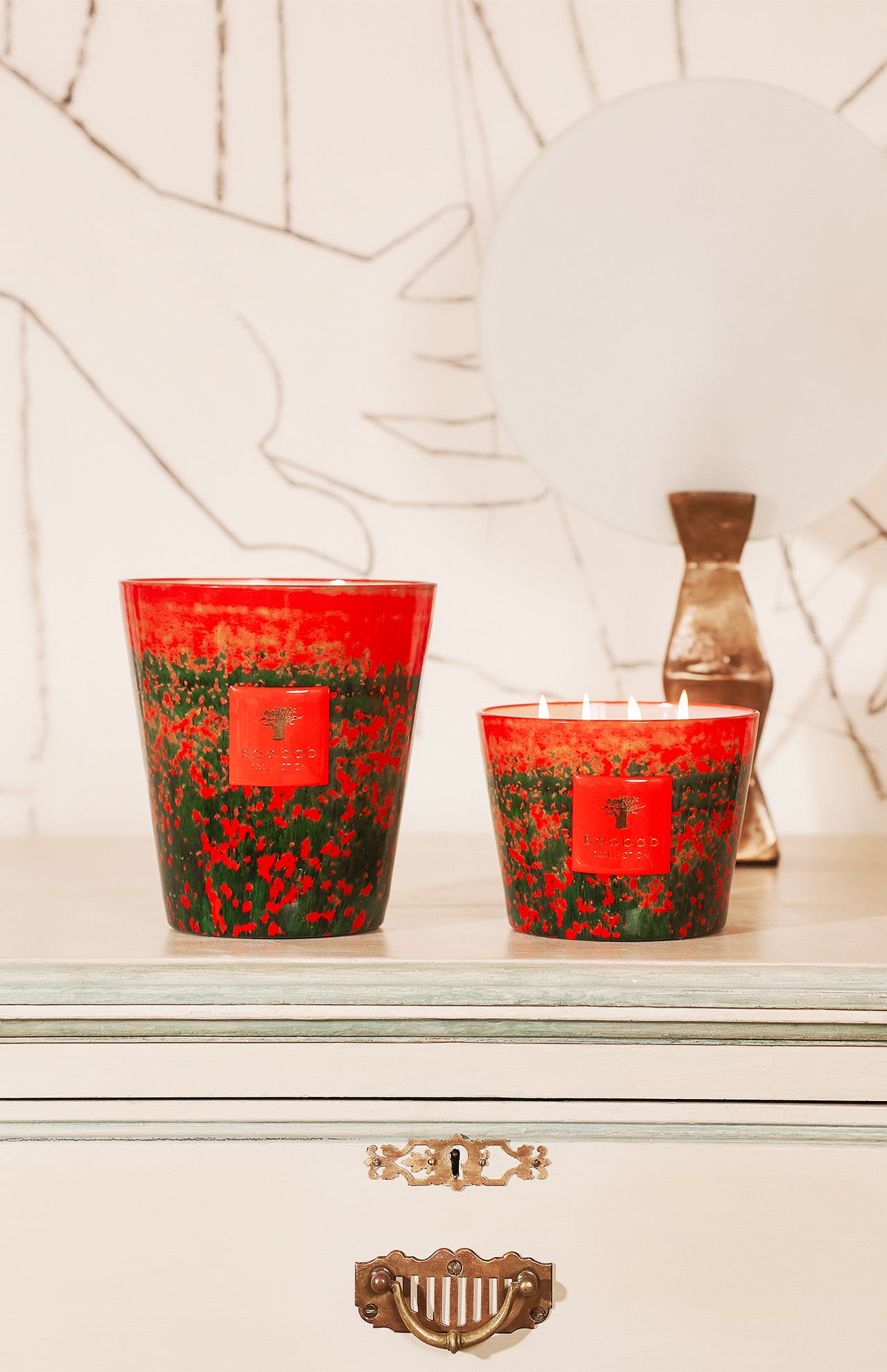 The BAOBAB COLLECTION Arty Flowers Poppies Max 16 Candle adds a decorative touch atop a light dresser, accompanied by a lamp with a round white shade and abstract wall art featuring a large hand outline in the background.
