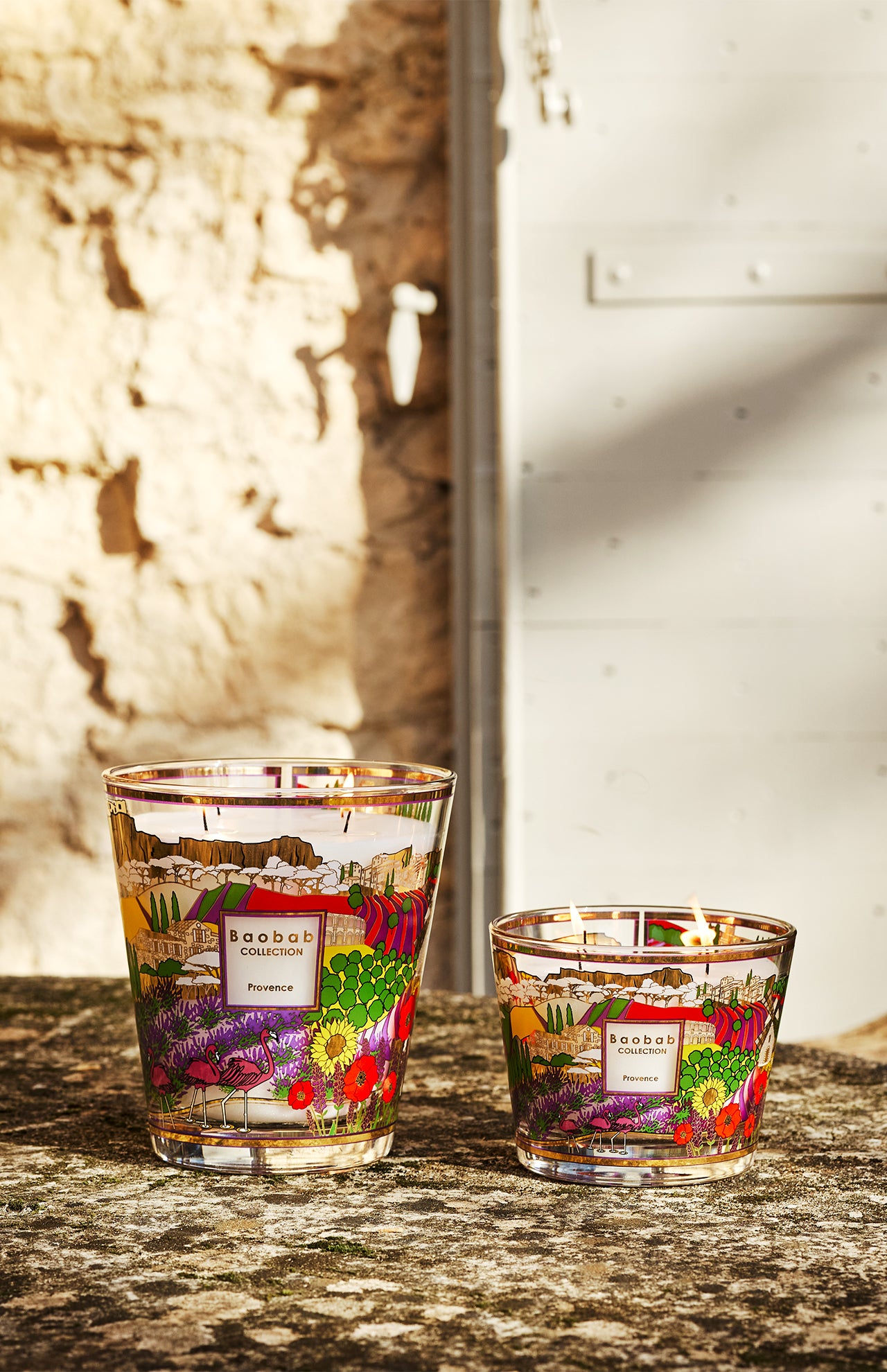 Two BAOBAB COLLECTION Provence Max 16 candles with vibrant floral patterns and hand-poured wax rest on a stone surface in sunlight, set against a textured beige wall and a white door.