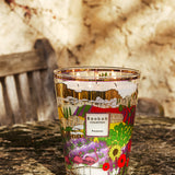 A BAOBAB COLLECTION Provence Max 24 Candle, decorated with vibrant floral and landscape designs, rests on a stone surface outdoors with a rustic bench and sunlit textured wall in the background.