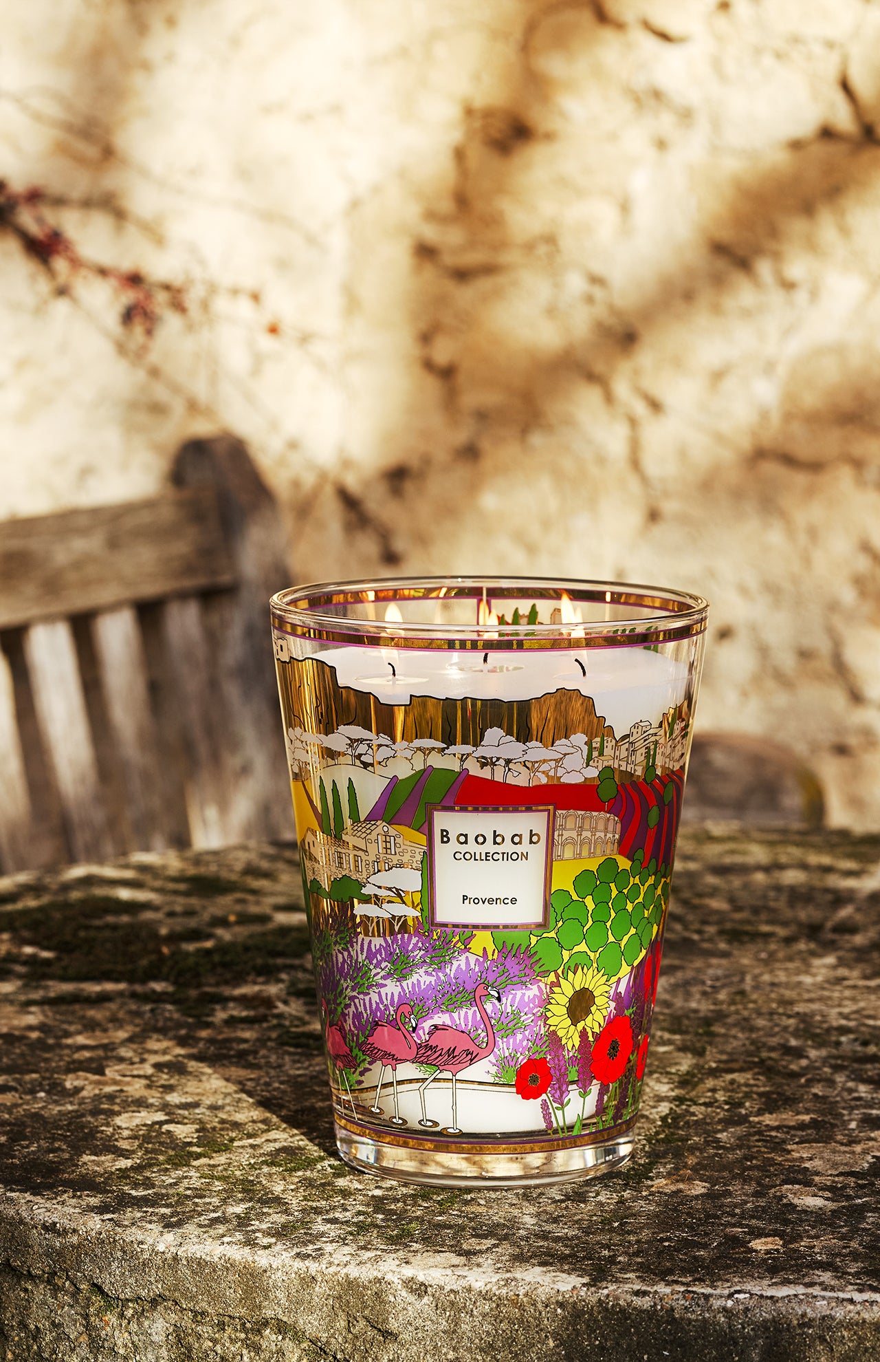 A BAOBAB COLLECTION Provence Max 24 Candle, decorated with vibrant floral and landscape designs, rests on a stone surface outdoors with a rustic bench and sunlit textured wall in the background.