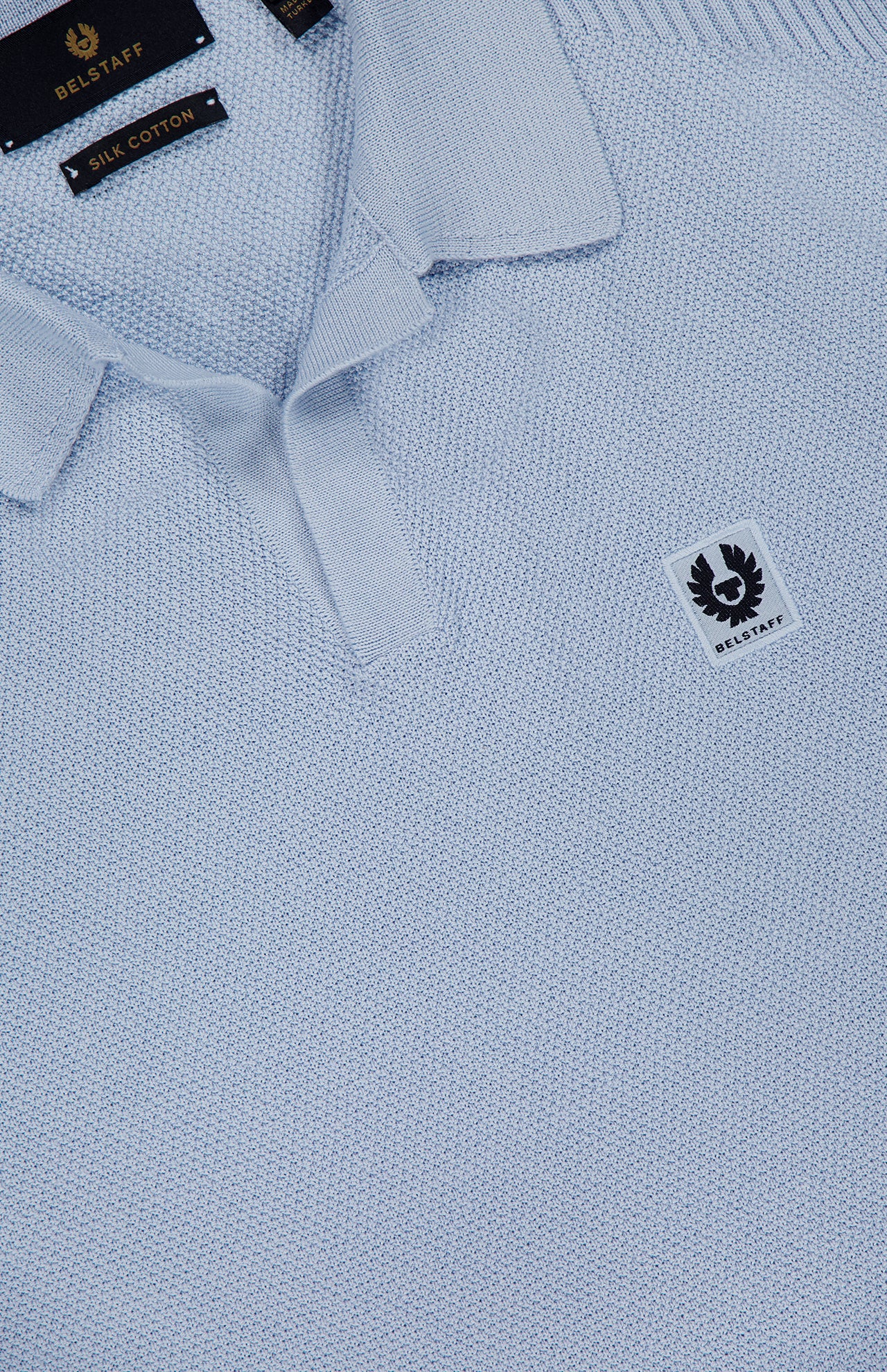 Close-up of the Coast Knitted Waffle Polo Shirt by BELSTAFF in light blue cotton silk piqué, featuring a textured knit, Johnny collar, short button placket, and a small chest patch with the brand logo. Inner label: Silk Cotton.
