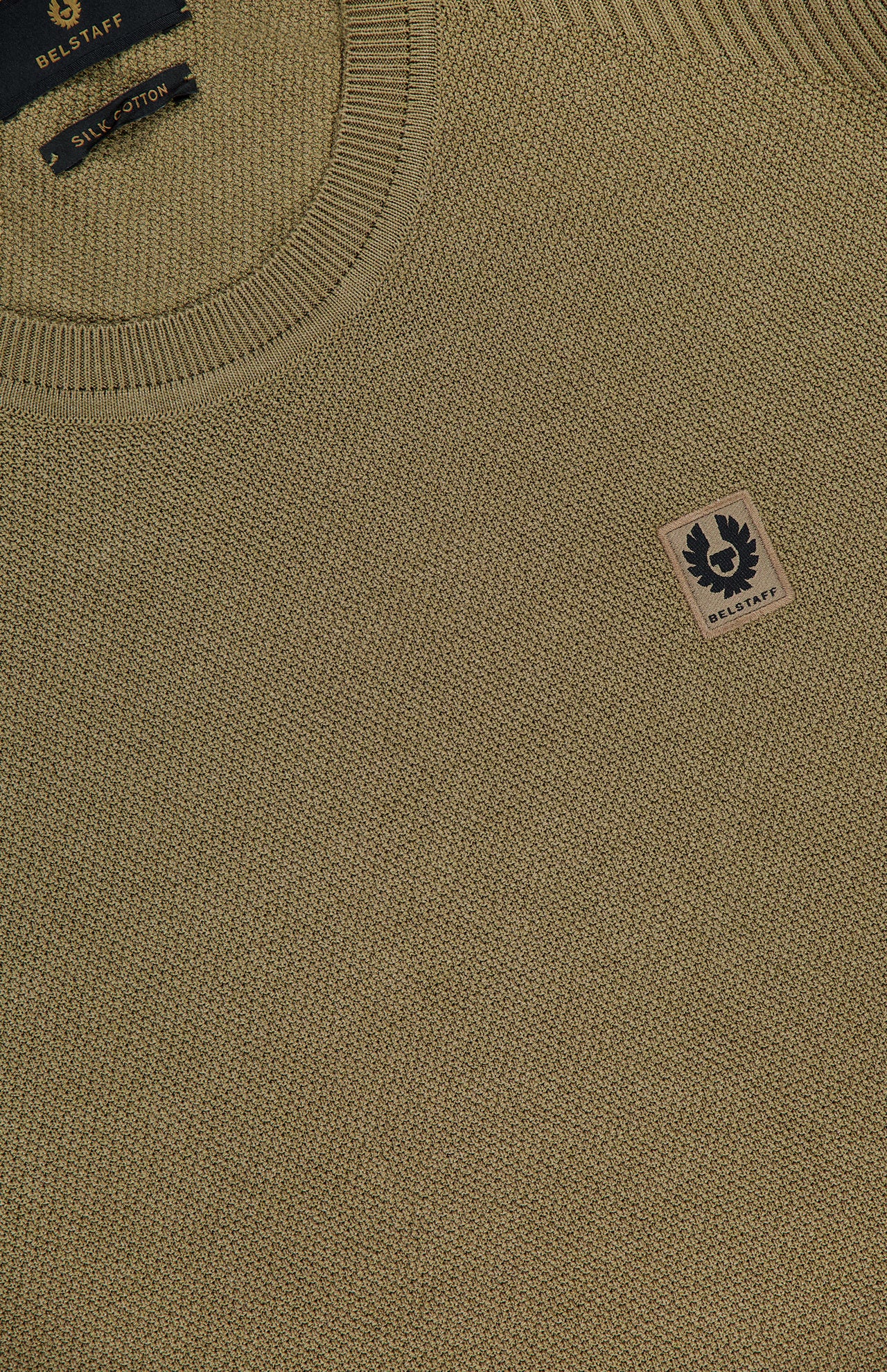 Close-up of a BELSTAFF Coast Knitted Sweater in olive green cotton-silk knit, featuring a ribbed crew neck, small beige logo patch with black emblem on the chest, and an inner label reading “Belstaff Silk Cotton.”.