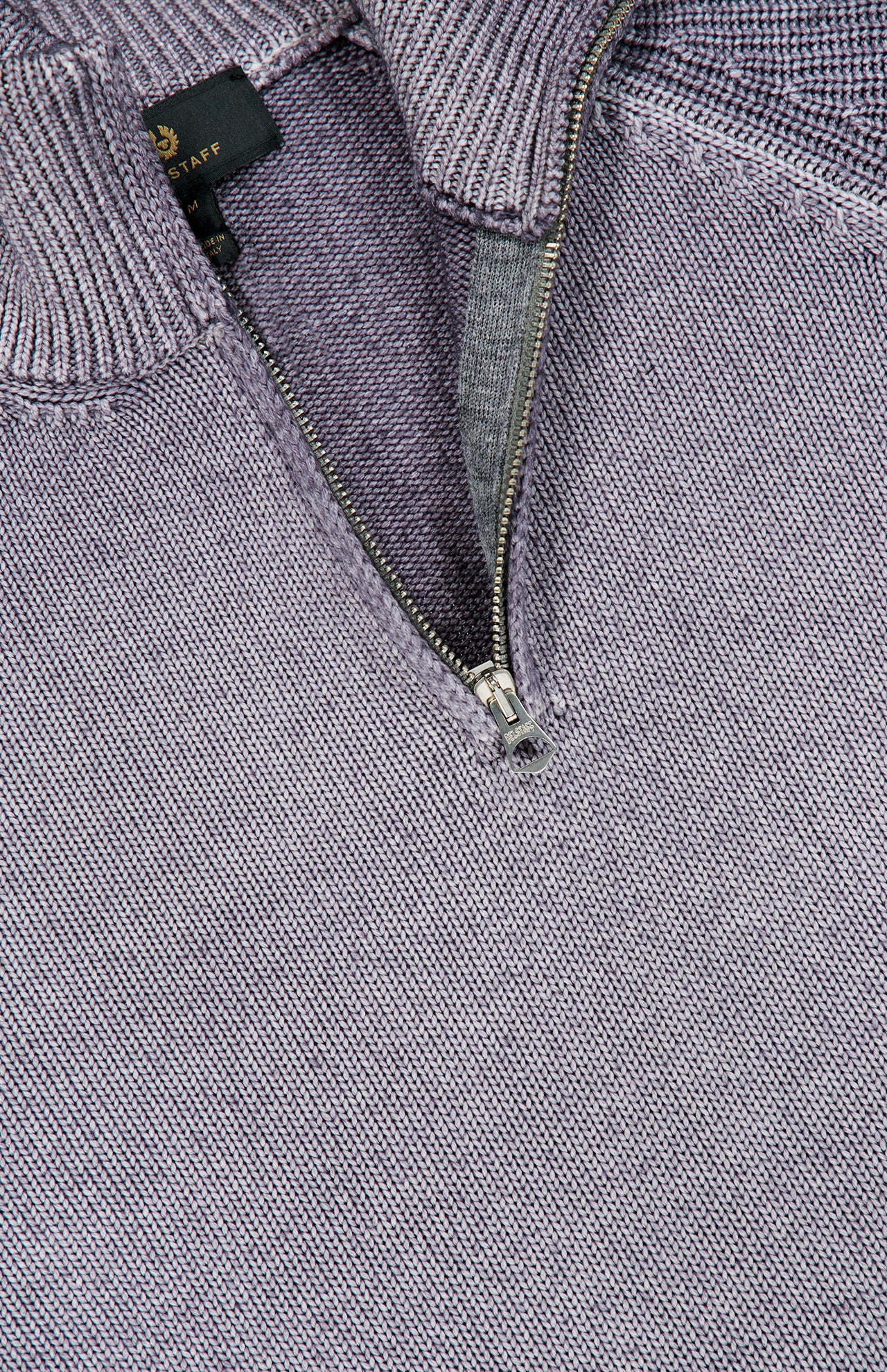 Close-up of the BELSTAFF Garment Dye Aspect Quarter Zip Sweater in light purple wool, featuring a partially unzipped metal quarter zip and ribbed collar with a small black tag visible inside the neckline. Made from responsibly-sourced wool for enhanced comfort and sustainability.