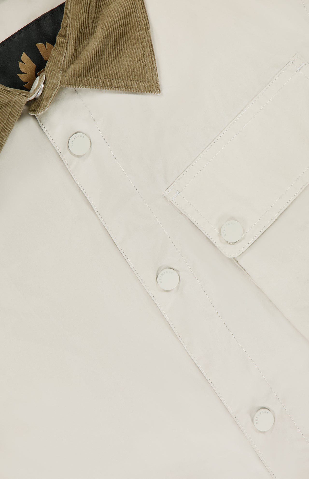 Close-up of the BELSTAFF Water Repellent Ground Jacket in cream, featuring a tan corduroy collar, white buttons, front chest pocket, and a visible black and gold inner label near the collar.