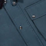 Close-up of the BELSTAFF Incline Field Jacket, featuring blue waxed cotton fabric with a smooth, slightly shiny finish that highlights its water- and wind-repellent qualities. The jacket showcases a corduroy collar, metal snap buttons, a flap pocket, and detailed stitching with precise button placement.