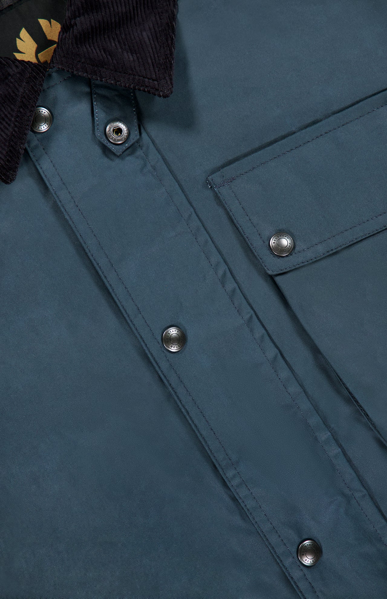 Close-up of the BELSTAFF Incline Field Jacket, featuring blue waxed cotton fabric with a smooth, slightly shiny finish that highlights its water- and wind-repellent qualities. The jacket showcases a corduroy collar, metal snap buttons, a flap pocket, and detailed stitching with precise button placement.