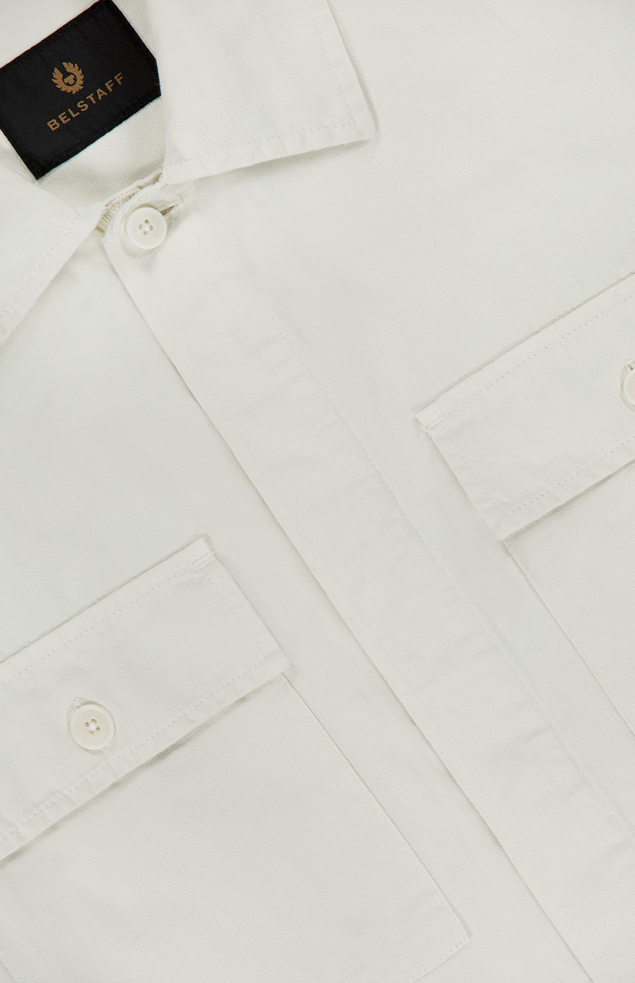 Close-up of the BELSTAFF Maritime Garment-dyed Overshirt in white cotton, featuring two buttoned chest pockets, buttoned front, part of the collar, and visible black and gold BELSTAFF tag—a stylish men's fashion staple.