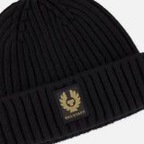 The BELSTAFF Watch Beanie Hat is a black ribbed knit wool beanie with a folded brim, adorned with a rectangular black and gold BELSTAFF logo patch on the front.