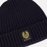 The BELSTAFF Watch Beanie Hat is crafted from dark navy ribbed knit lambswool and features a rectangular patch with the brand’s gold logo and text on the front. The hat is shown against a plain white background.