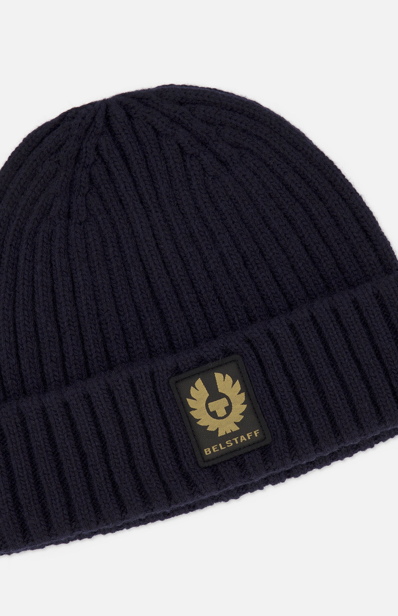 The BELSTAFF Watch Beanie Hat is crafted from dark navy ribbed knit lambswool and features a rectangular patch with the brand’s gold logo and text on the front. The hat is shown against a plain white background.