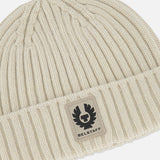 A close-up of the BELSTAFF Watch Beanie Hat, crafted from cream-colored ribbed knit lambswool with a folded cuff and finished with a rectangular Belstaff logo patch on the front—an ideal addition to your cold-weather accessories collection.