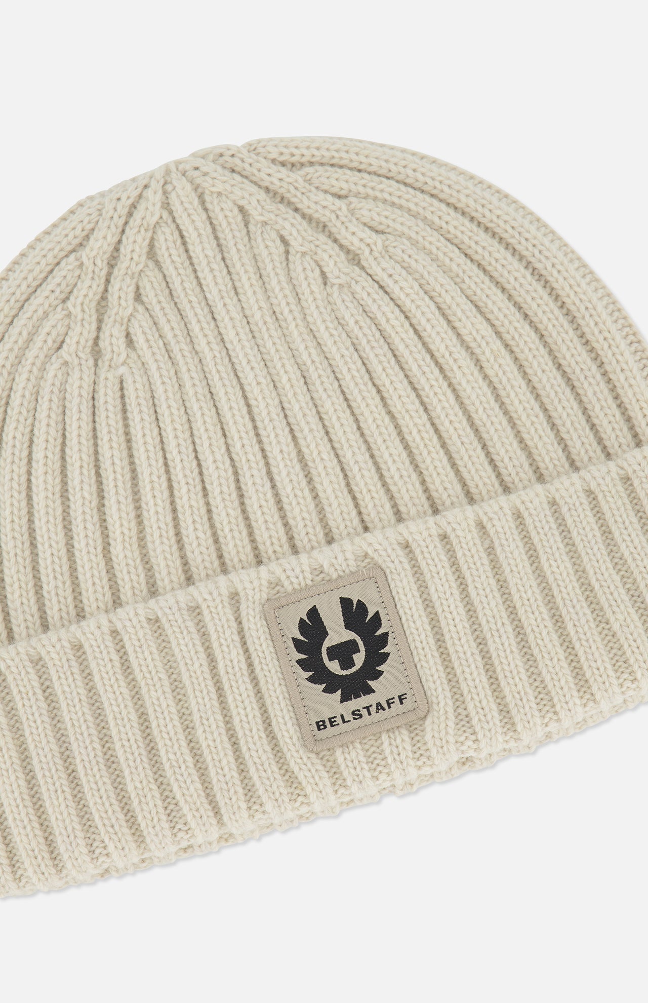 A close-up of the BELSTAFF Watch Beanie Hat, crafted from cream-colored ribbed knit lambswool with a folded cuff and finished with a rectangular Belstaff logo patch on the front—an ideal addition to your cold-weather accessories collection.