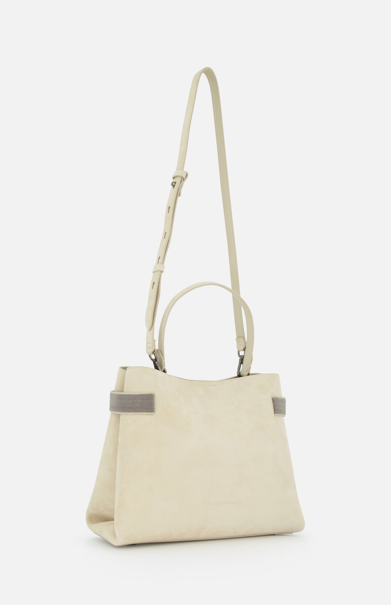 The Medium Soft Velour Essence Bag by BRUNELLO CUCINELLI features beige suede, two short handles, an adjustable shoulder strap, and gray-edged handles that highlight its minimalist design and structured shape against a white background.