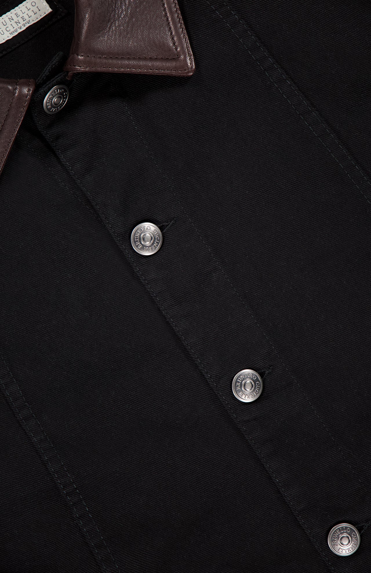 Close-up of the BRUNELLO CUCINELLI Denim with Leather Collar Jacket, featuring black denim, silver buttons, a brown leather collar, and a visible clothing label—showcasing timeless retro style.