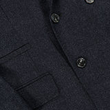Close-up of the BRUNELLO CUCINELLI Double Breasted Suit Jacket, crafted from dark, textured virgin wool. The jacket features overlapping lapels, a visible pocket, and glossy black buttons that add a subtle sheen to the refined fabric.