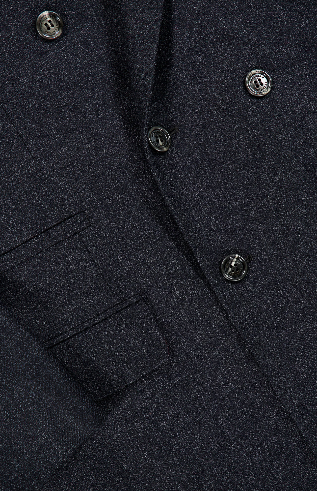 Close-up of the BRUNELLO CUCINELLI Double Breasted Suit Jacket, crafted from dark, textured virgin wool. The jacket features overlapping lapels, a visible pocket, and glossy black buttons that add a subtle sheen to the refined fabric.