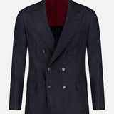 The BRUNELLO CUCINELLI Double Breasted Suit Jacket is shown on a plain white background. This dark navy jacket features six buttons, peaked lapels, and two front flap pockets. Expertly tailored from virgin wool, it has a luxurious deep burgundy interior lining.
