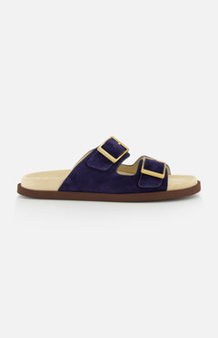 The BRUNELLO CUCINELLI Double Buckle Suede Sandals are slip-ons in navy with two wide straps, large gold buckles, a light beige footbed, and a flat brown sole, pictured in profile on a white background.