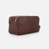 The BRUNELLO CUCINELLI Leather Dopp Kit, expertly crafted in Italy, is one of the finest travel accessories. Made from brown leather, it features a zippered top and a small handle on one end, and is displayed against a plain white background.