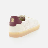 The BRUNELLO CUCINELLI Suede Leather Detailing Sneakers feature a cream-colored suede upper, white laces, tan gum sole, and a burgundy heel tab with a gold lion logo, shown from a rear side angle on a white background.