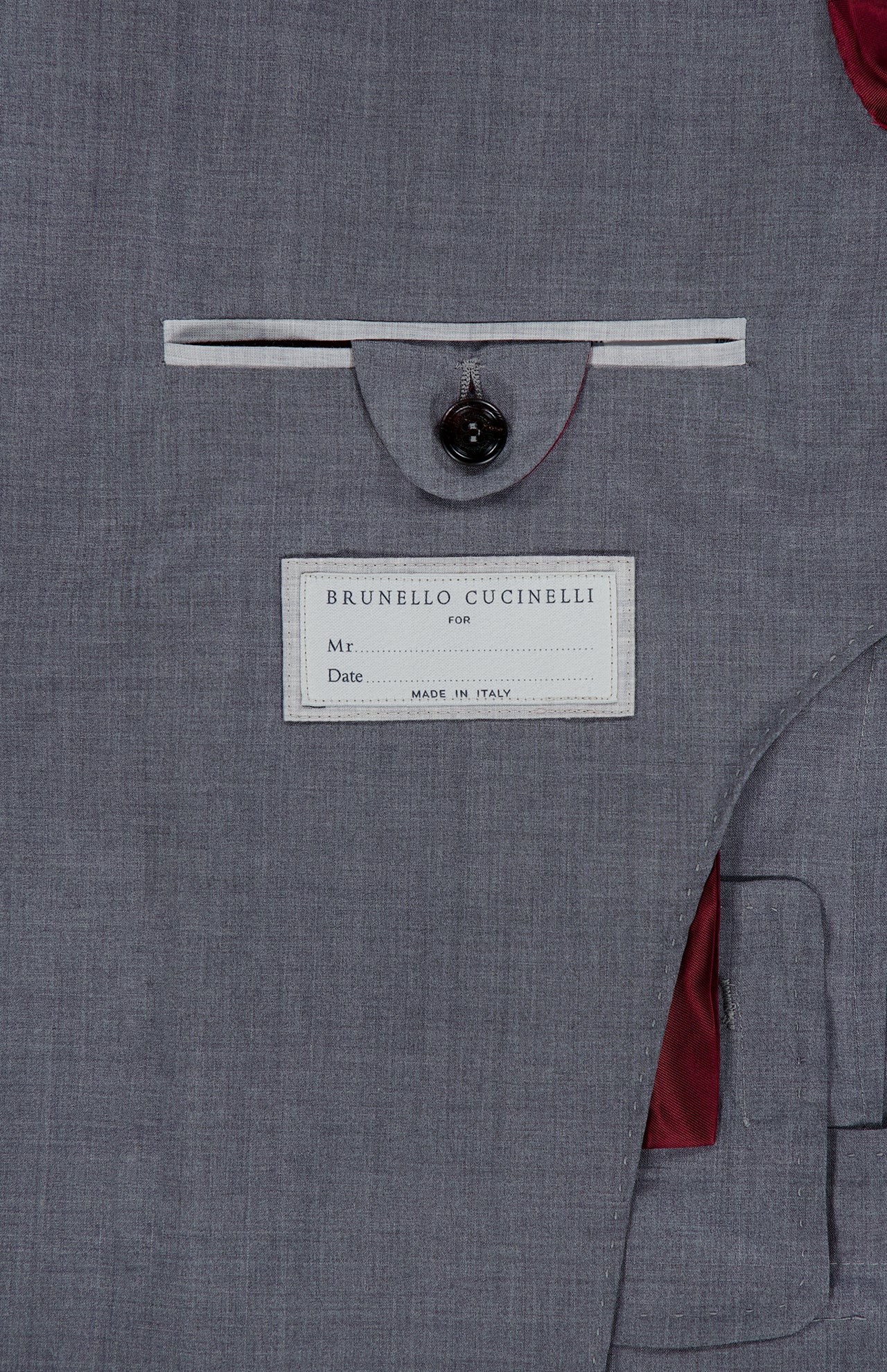 Detailed close-up of the inner pocket on the grey BRUNELLO CUCINELLI Wool and Silk Travel Blazer, showcasing a button closure and a Brunello Cucinelli label with spaces for “Mr.” and “Date,” along with the inscription “Made in Italy.” The partially visible lining is red.