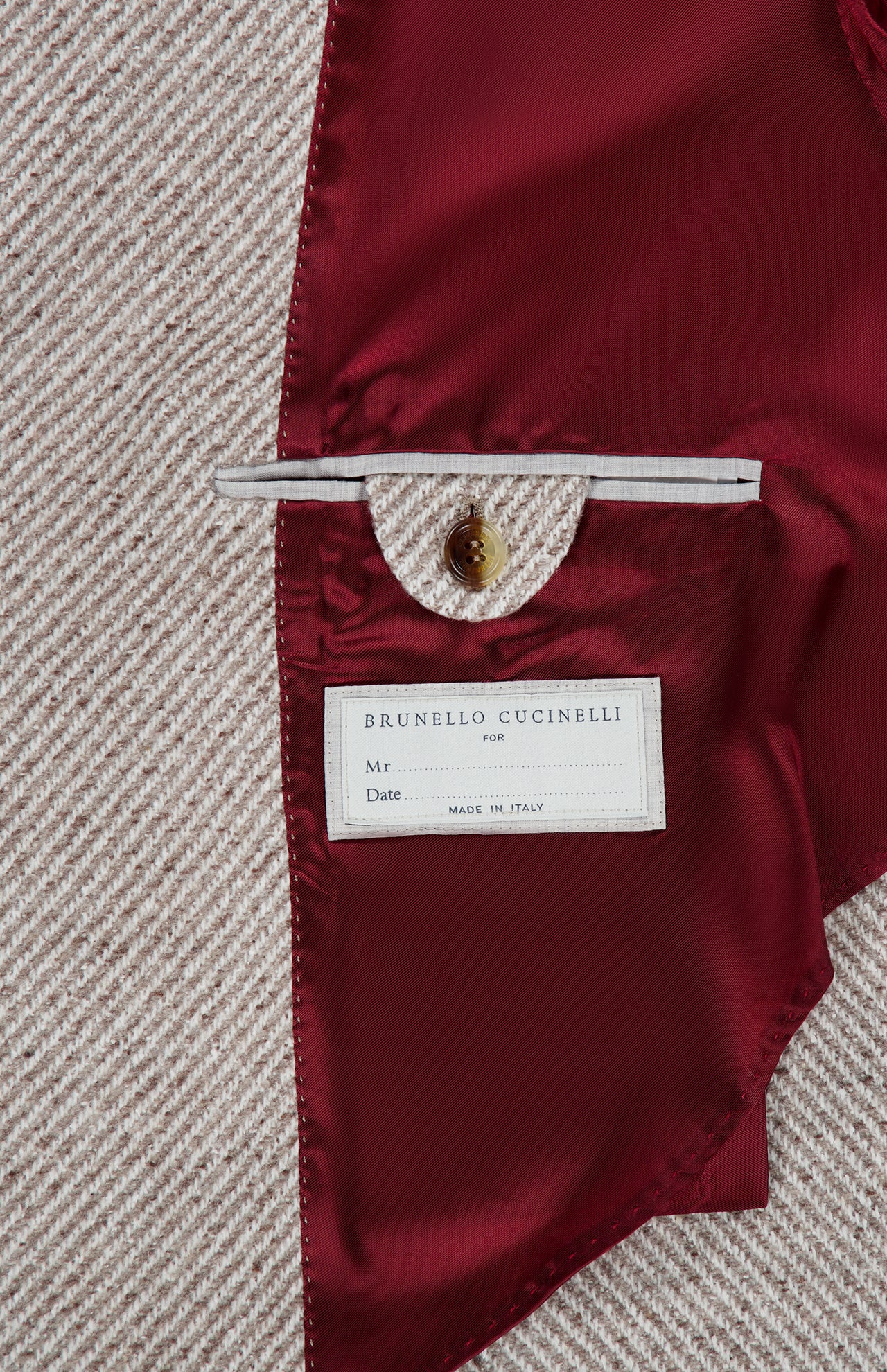 Close-up of the Water Resistant Cashmere Sport Jacket’s interior reveals a luxurious red satin lining, a buttoned pocket, and a white BRUNELLO CUCINELLI label with space for personal details, set against beige textured fabric. Exquisitely crafted in Italy by BRUNELLO CUCINELLI, this jacket exemplifies timeless sophistication.