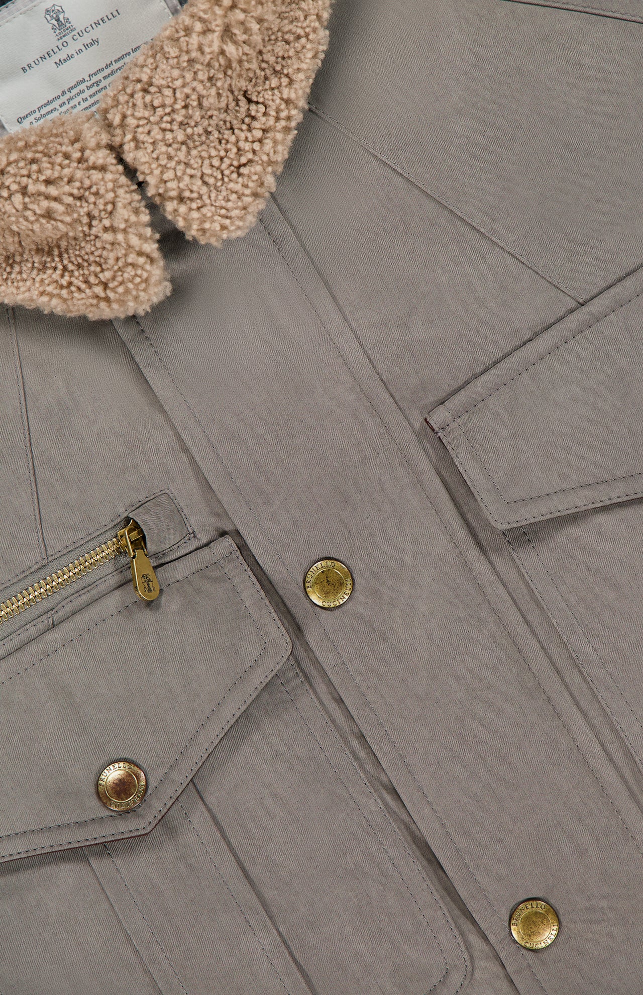 Close-up of the BRUNELLO CUCINELLI Water Resistant Safari Jacket in taupe, featuring gold buttons, a gold-zippered pocket, flap pockets, and a tan sherpa collar. The interior displays a white "Brunello Cucinelli Made in Italy" label.