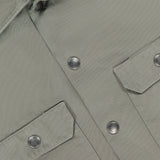 Close-up of the BRUNELLO CUCINELLI Western Cotton Long Sleeve Shirt in light olive green, crafted from garment-dyed gabardine cotton and featuring two front flap pockets, metal snap buttons, and a visible white label at the collar.