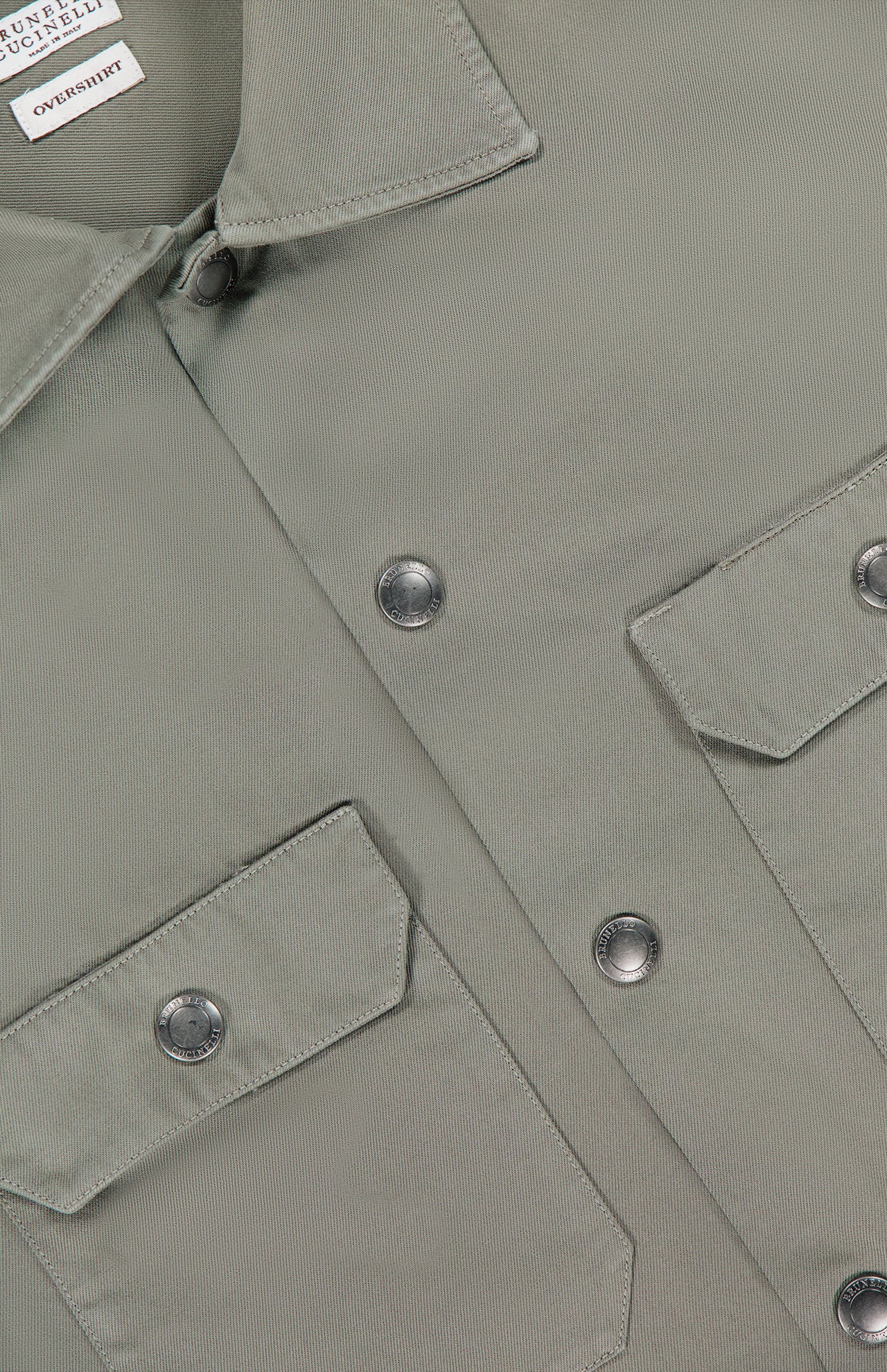 Close-up of the BRUNELLO CUCINELLI Western Cotton Long Sleeve Shirt in light olive green, crafted from garment-dyed gabardine cotton and featuring two front flap pockets, metal snap buttons, and a visible white label at the collar.