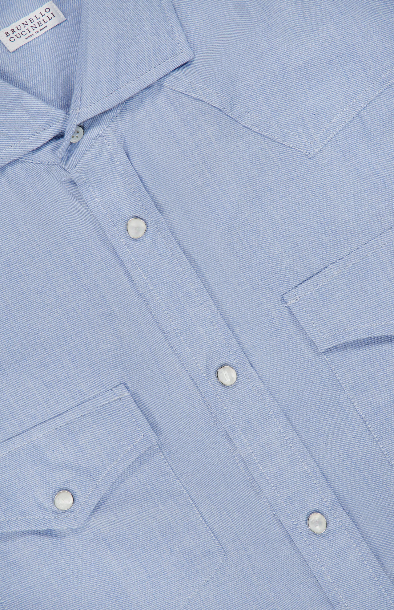Close-up of the BRUNELLO CUCINELLI Cotton and Cashmere Western Shirt in light blue, featuring two chest pockets and pearl snap buttons for classic western wear appeal. The subtly textured fabric is highlighted, with a white BRUNELLO CUCINELLI label visible inside the collar.