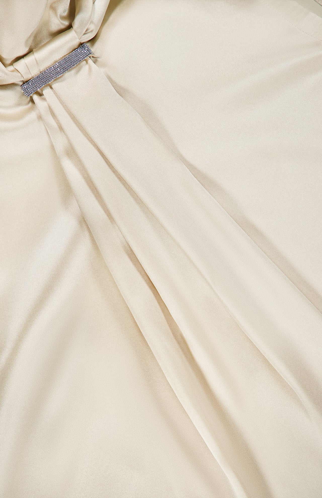Close-up of a smooth, cream-colored silk satin fabric with gentle folds and a rhinestone-embellished buckle detail near the top left corner, evocative of the luxurious finish on the BRUNELLO CUCINELLI Silk Satin Tie Sleeveless Blouse.