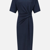 BRUNELLO CUCINELLI’s Wool Jersey Wrap Dress in navy features a knee-length silhouette with short sleeves, a high neckline, and a gathered twist detail at the waist. Simple yet refined, this elegant dress is displayed against a plain white background.