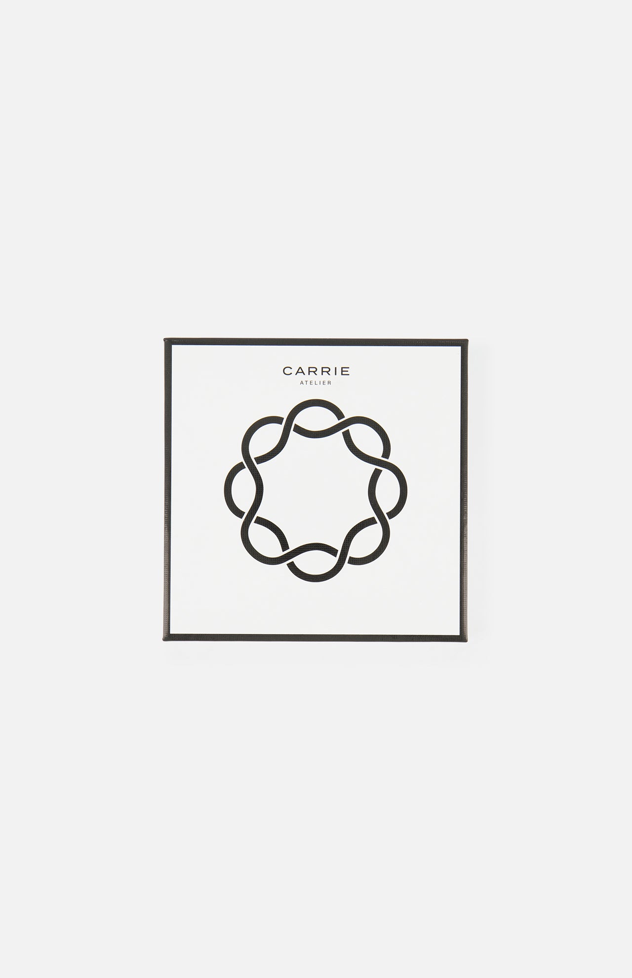 A white square box with a black abstract circle in the center and “CARRIE” above—stylish packaging for the Carrie Purse Hook Suede Etoile in Rose Eclipse by CARRIE ATELIER, a chic designer bag accessory.