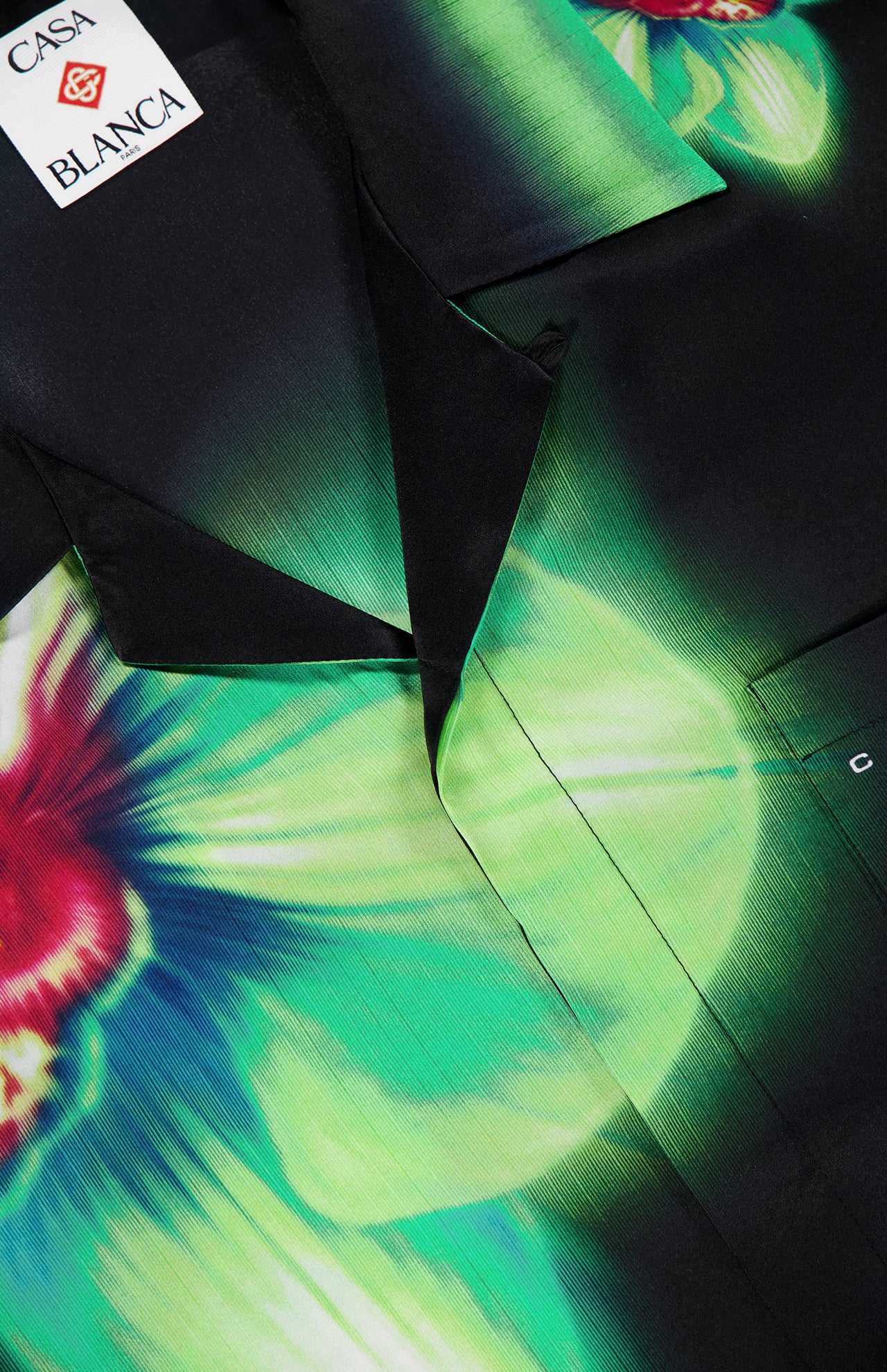 Close-up of the CASABLANCA Glitch Orchid Cuban Collar Short Sleeve Shirt featuring a bold green and red orchid print on silky black fabric, plus the signature label sewn inside the neckline for a luxurious finish.