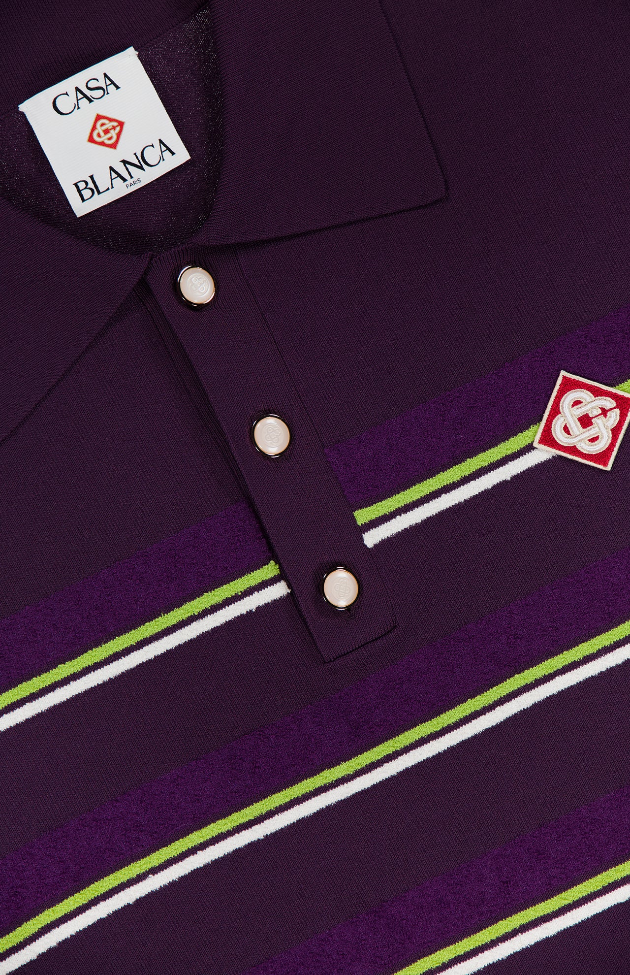 Close-up of the CASABLANCA Stripe Short Sleeve Polo Shirt in dark purple, showcasing green and white stripes, three metallic buttons, and an embroidered red and white logo patch on the chest.