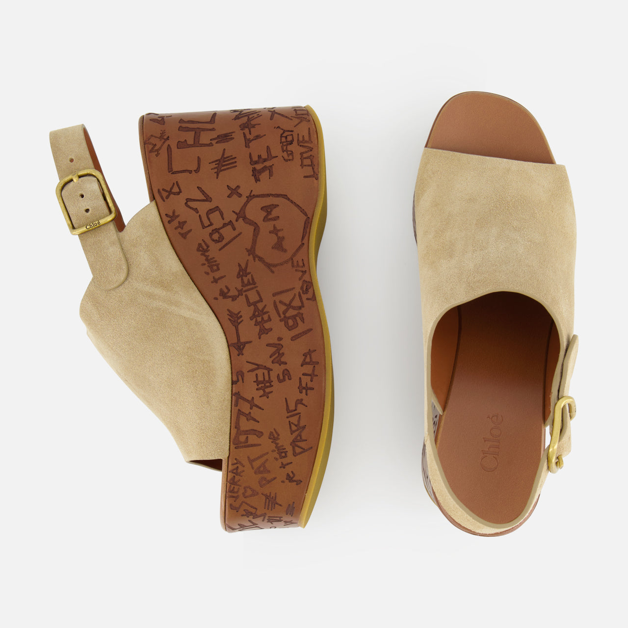 The CHLOÉ Maxime Carved Wedge Heel features tan suede clogs with open toes, gold buckles, brown platform soles adorned with graffiti-style markings and text, and a bold wood-effect wedge heel.