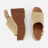 The CHLOÉ Maxime Carved Wedge Heel features tan suede clogs with open toes, gold buckles, brown platform soles adorned with graffiti-style markings and text, and a bold wood-effect wedge heel.