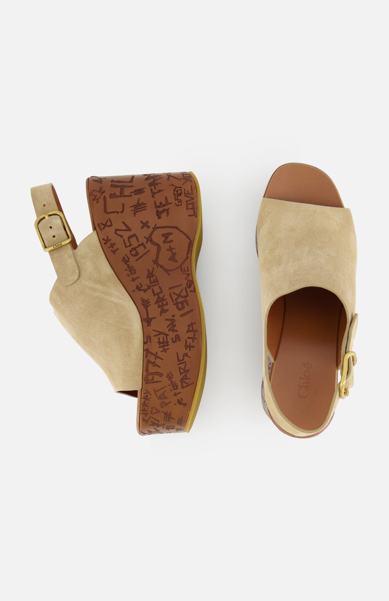 The CHLOÉ Maxime Carved Wedge Heel features tan suede clogs with open toes, gold buckles, brown platform soles adorned with graffiti-style markings and text, and a bold wood-effect wedge heel.