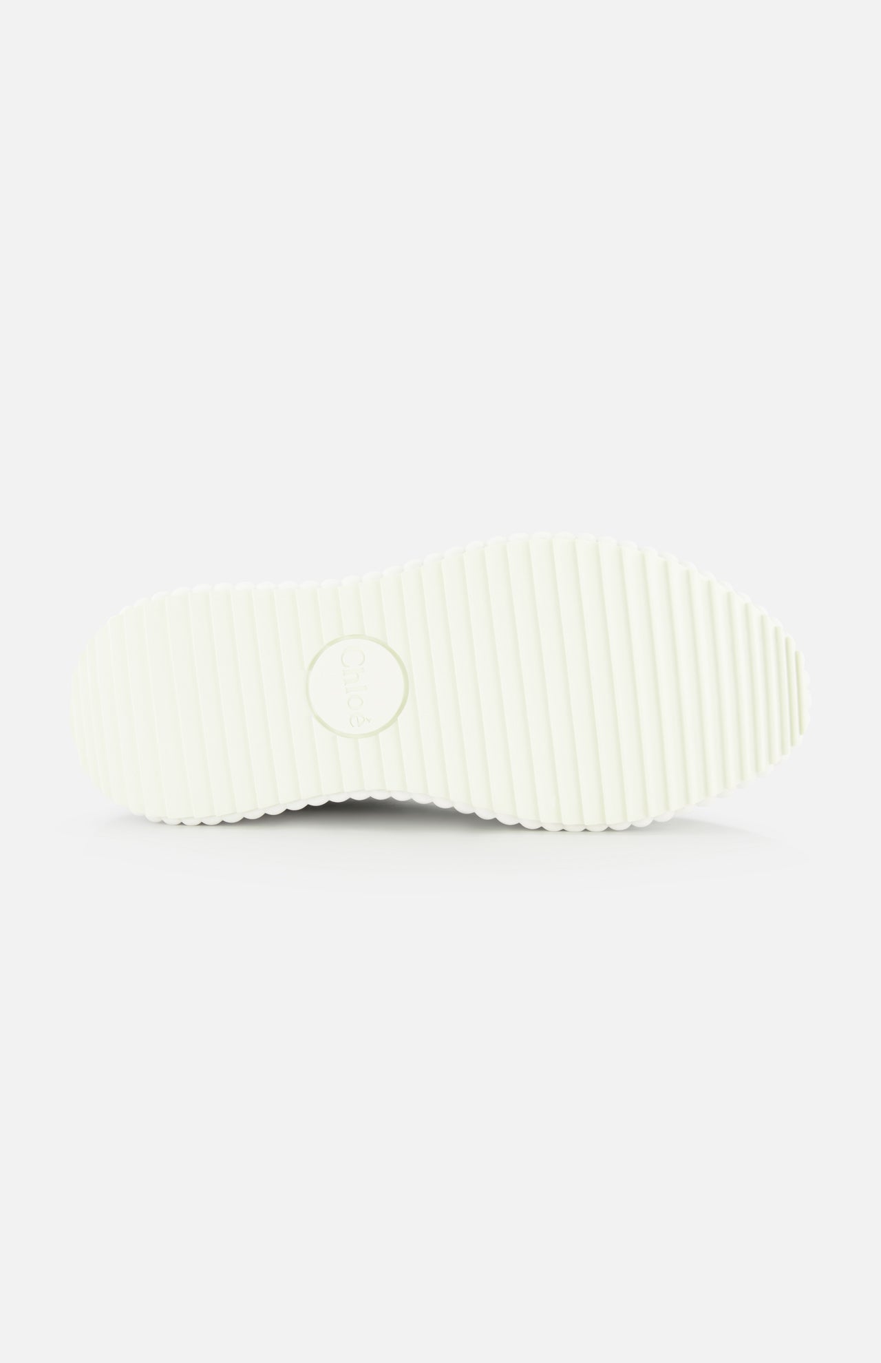 The image shows the bottom view of CHLOÉ Metallized Nama Sneakers' white sole with a ridged, grooved texture and a circular logo at the center, highlighting its recycled sole against a plain white background.