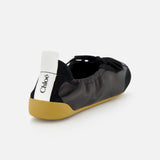 The CHLOÉ Laced Kick Flats feature black leather, a tan rubber sole, a black bow on top, and a white pull tab with the Chloé logo. These stylish slip-ons are shown against a plain white background.