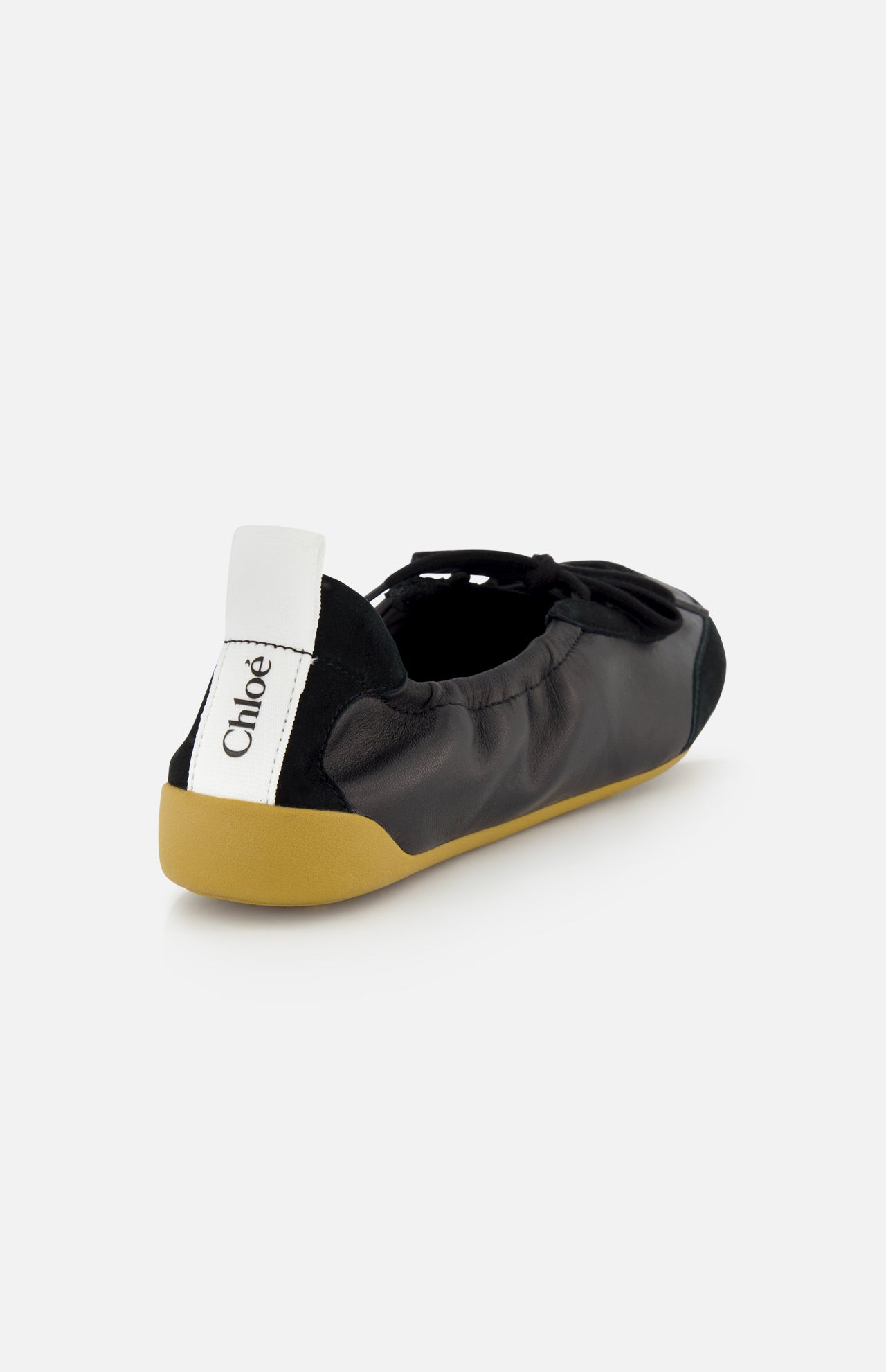The CHLOÉ Laced Kick Flats feature black leather, a tan rubber sole, a black bow on top, and a white pull tab with the Chloé logo. These stylish slip-ons are shown against a plain white background.