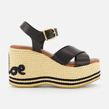 The CHLOÉ Chlóe Sand Wedge Heel is a black leather sandal with crisscross straps, ankle buckle, espadrille-style jute sole with black stitching, wavy heel design, and classic wedge heel flair.
